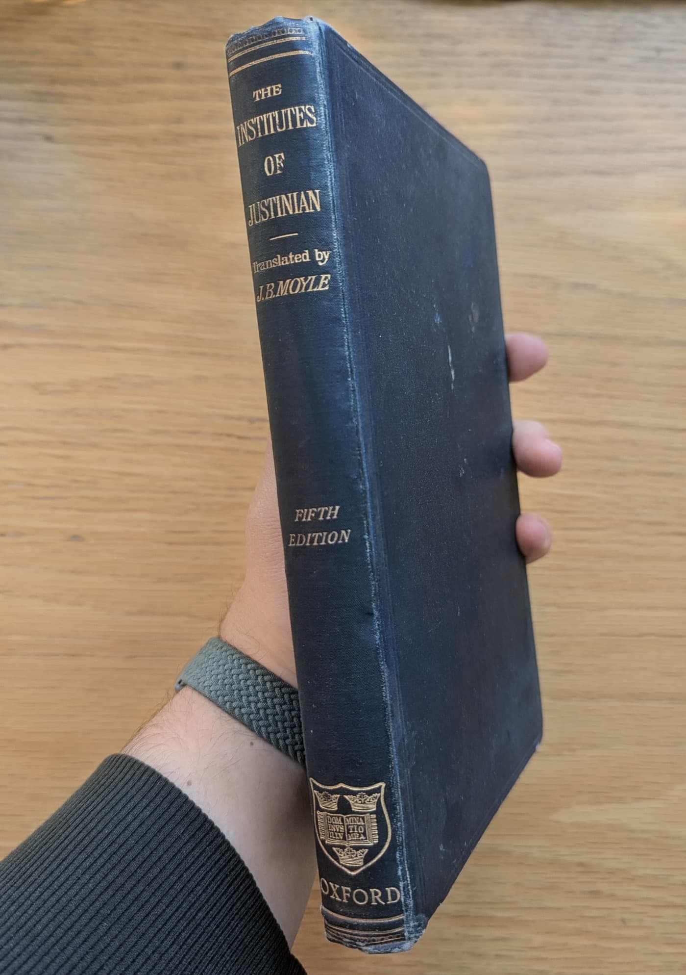 A hand holding the Institutes of Justinian, translated by J. B. Moyle, fifth edition, Oxford, dark blue cloth binding with gilt spine